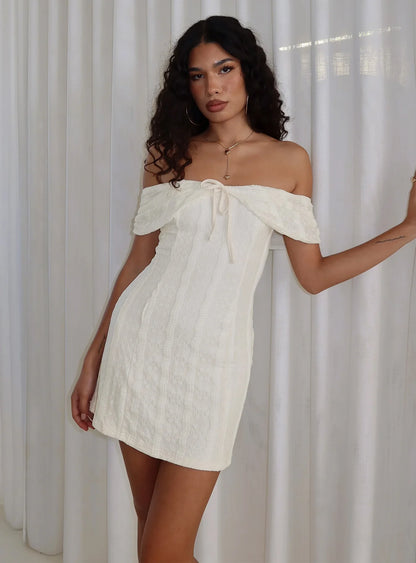 Model wearing a cream off-shoulder mini homecoming dress with bow detail.
