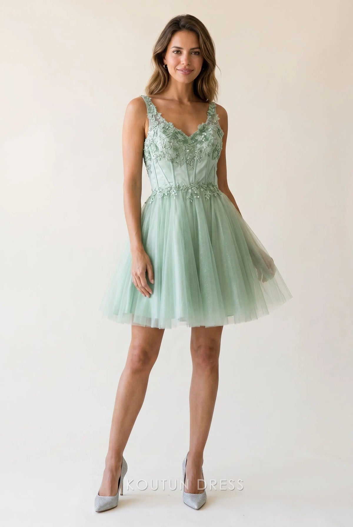 Model wearing a sage green glitter tulle mini homecoming dress with a V-neck and floral lace applique.
